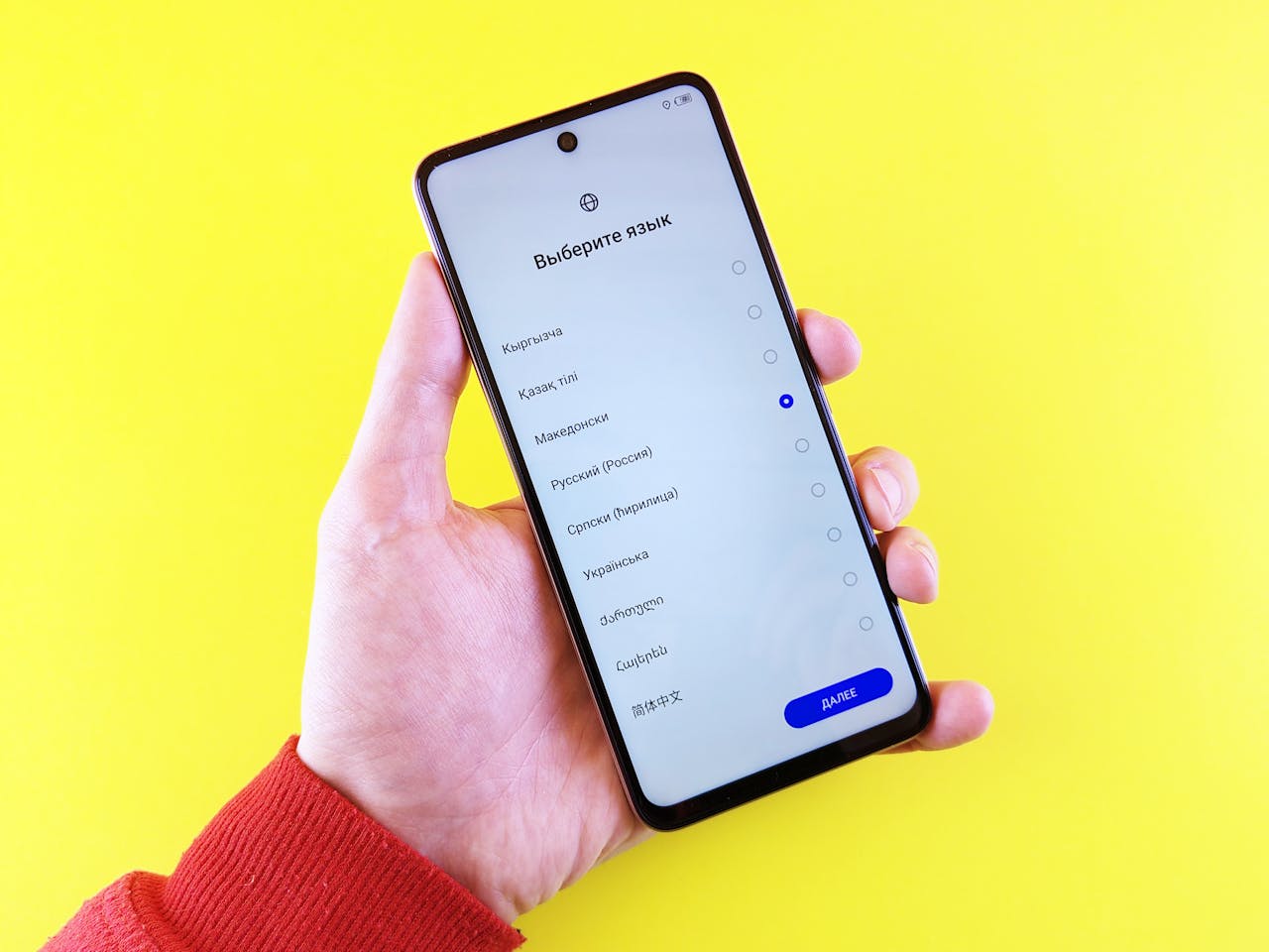 A hand in a red sleeve holding a smartphone displaying a language selection screen against a yellow background.